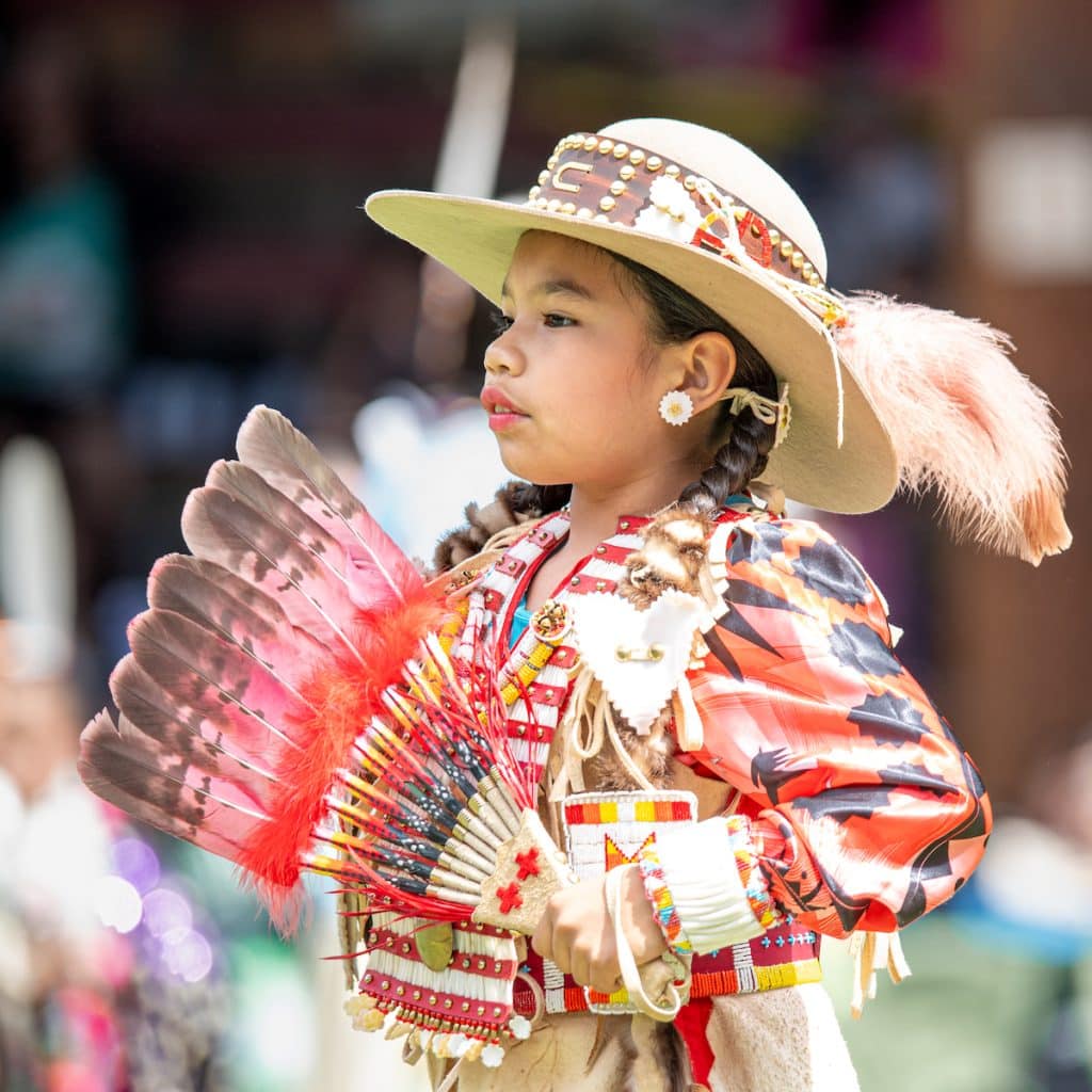 A young dancer wears regalia, long braids wrapped in otter furs and a feathered hat.