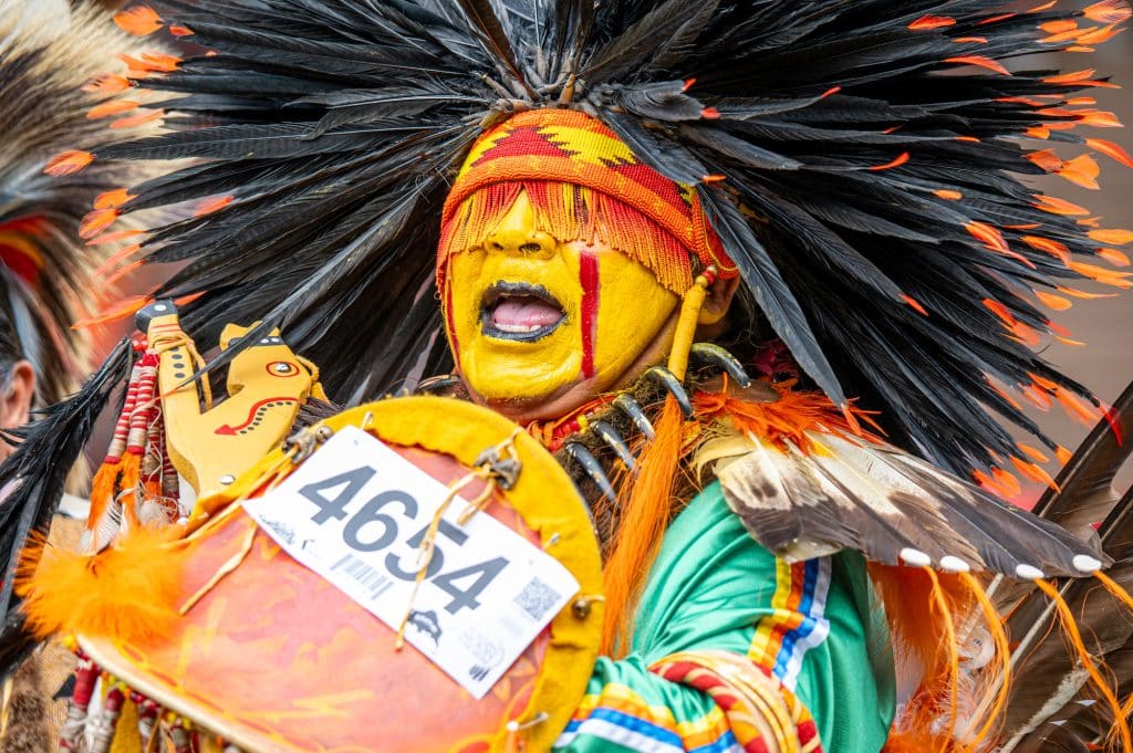 A portrait image shows powwow dancer Elvin Nicoteen wearing yellow paint and a feather hat.