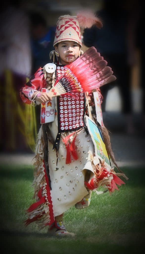 A young dancer wears elk regalia with long braids wrapped in otter fur, adorned with soft pink conch shells that shimmered under the sunlights. She wore an elk tooth buckskin dress and moccasins with her basket hat that speak to the beauty and strength of her St’át’imc and Warm Springs heritage.