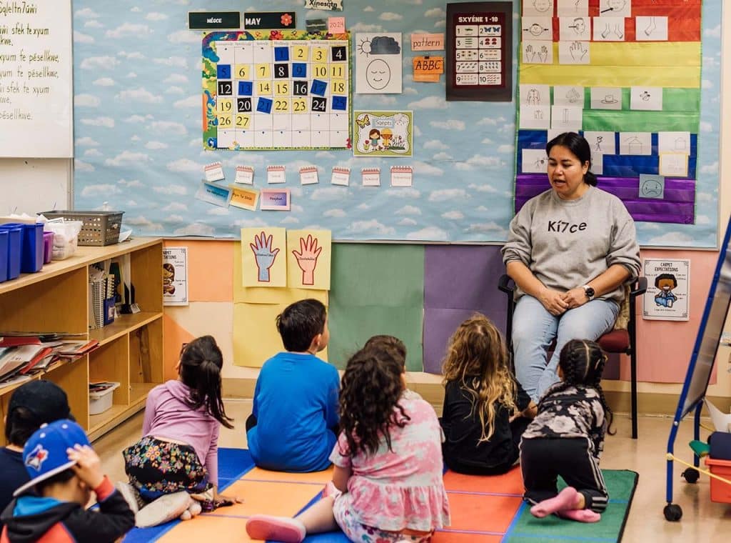 A teacher sits with a group of kids on the floor listening in a classroom with bright colours 