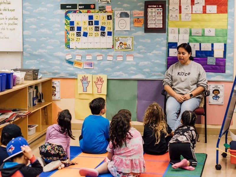 A teacher sits with a group of kids on the floor listening in a classroom with bright colours