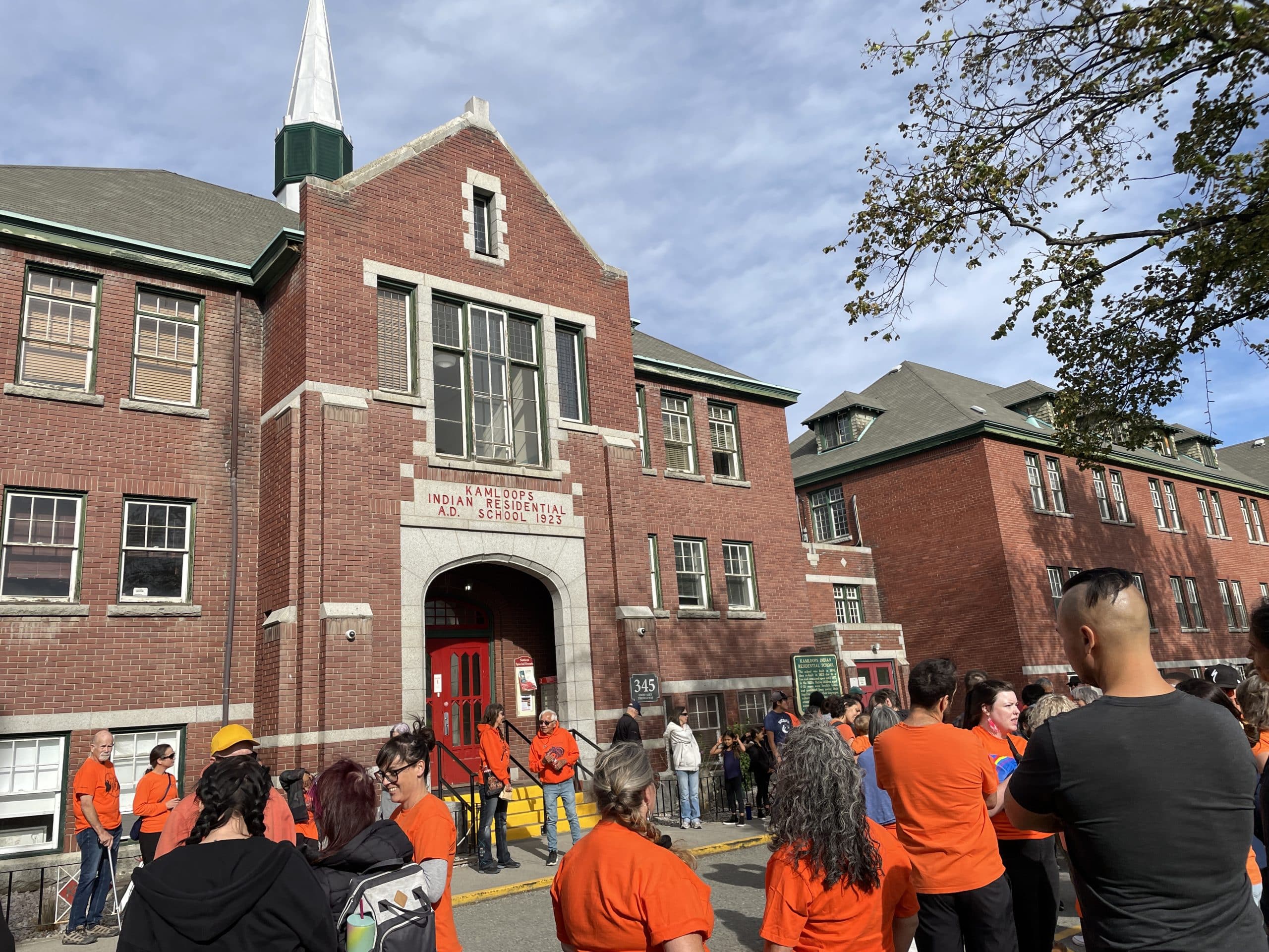 People dressed in orange stand outside a brick building for Orange Shirt Day.