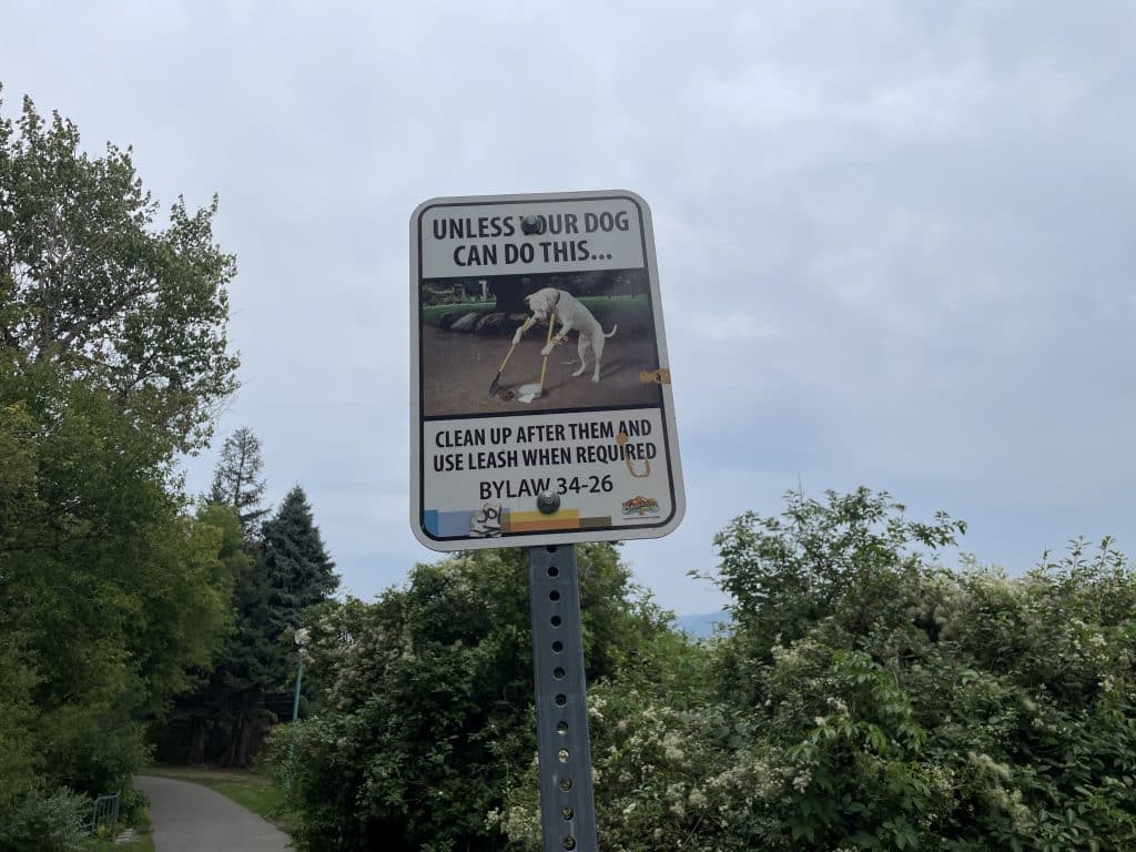 A sign states "Unless your dog can do this... [the image shows a dog picking up poo from the ground with a broom and dustpan] clean up after them and use leash when required. Bylaw 34-26.
Dog poop in Kamloops 