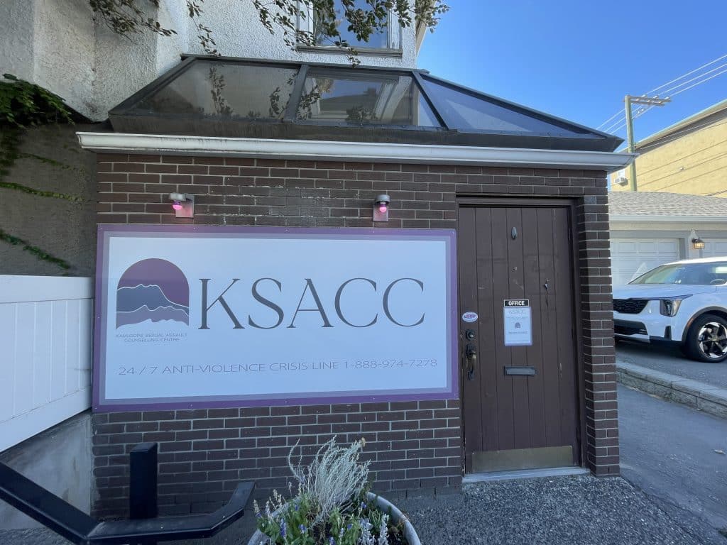 A photo of the office of the Kamloops Sexual Assault Counselling Centre. It has a big white poster with the letters KSACC and smaller words below which show what it is. This is the entrance to the office. 