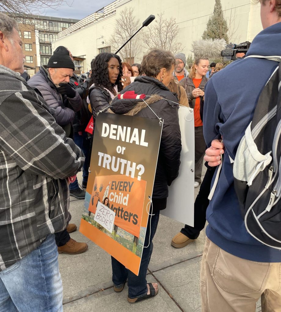 Various individuals gathered around a women with a sign that reads "Denial or Truth?" which contributes to the residential school denialism