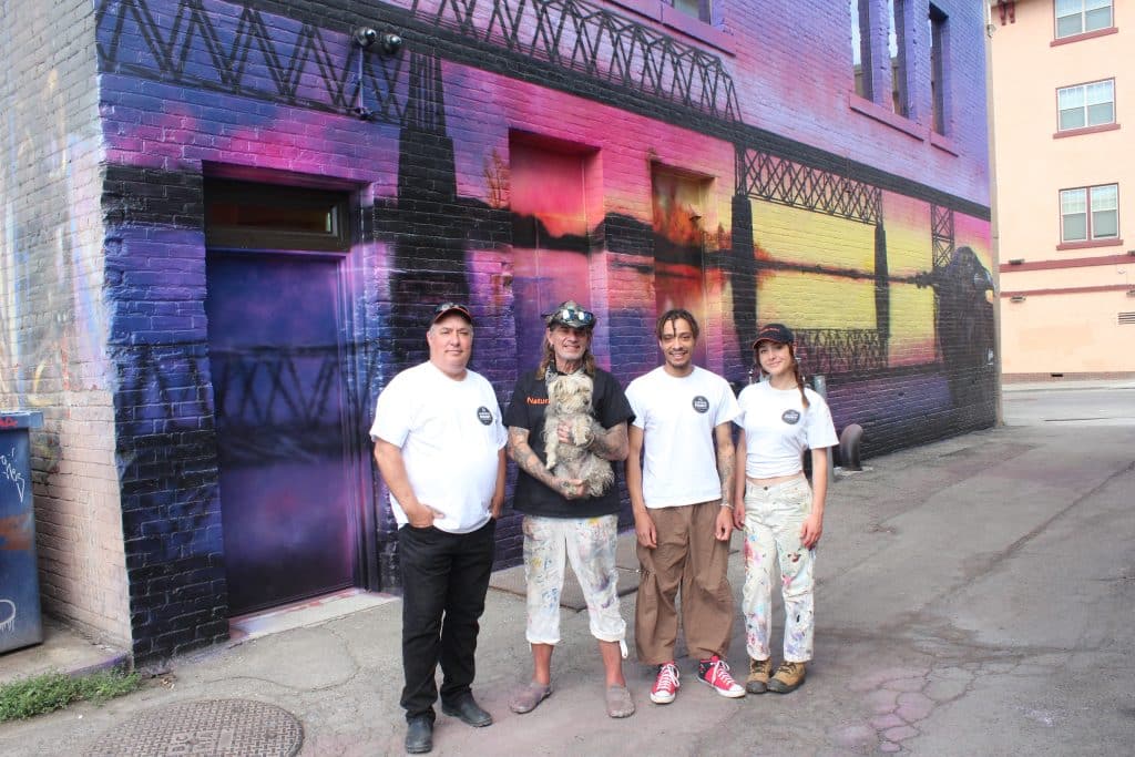 Vaughn Warren and his mentor, Paul Archer, stands with two young artists, Rowan Hunt and Jayde Richter, from Old Federal Studios, outside of a mural with vibrant sunset colours of the old Red Bridge