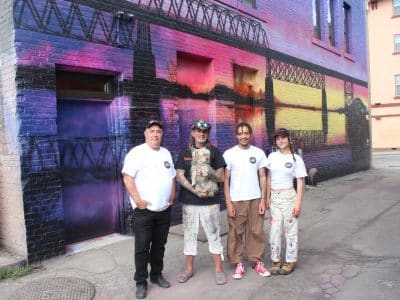 Vaughn Warren and his mentor, Paul Archer, stands with two young artists, Rowan Hunt and Jayde Richter, from Old Federal Studios, outside of a mural with vibrant sunset colours of the old Red Bridge