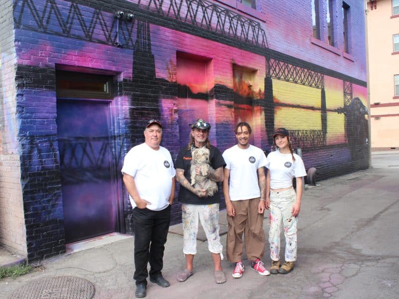 Vaughn Warren and his mentor, Paul Archer, stands with two young artists, Rowan Hunt and Jayde Richter, from Old Federal Studios, outside of a mural with vibrant sunset colours of the old Red Bridge