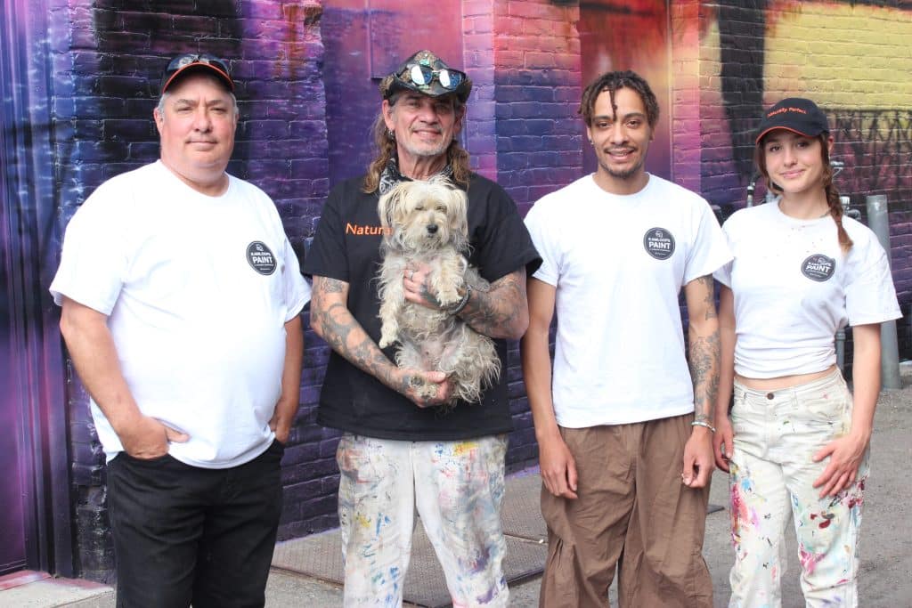 A mural team of four pose in front of the purple and pink mural wall in the back alley downtown