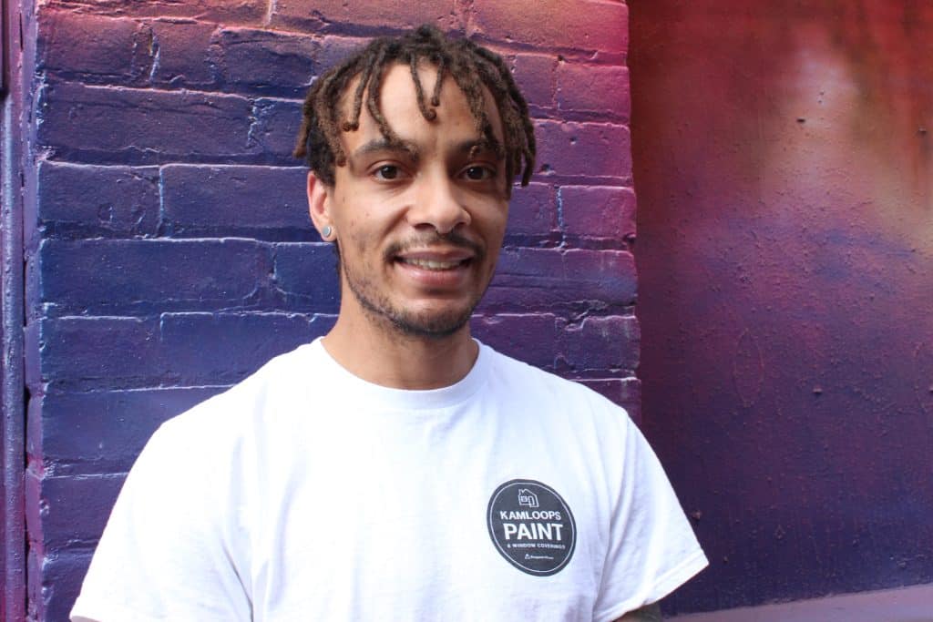 A man with short dreads smiles for the camera wearing a bright white t shirt and mural in background